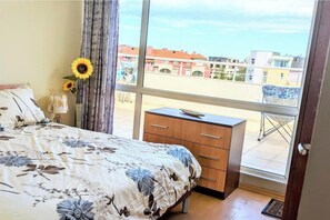 Ein Schlafzimmer mit Bett, Nachttisch, Kommode und Fenster mit Blick auf Gebäude.