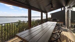 Une terrasse extérieure couverte, dotée d’une longue table et de chaises, qui offre une vue sur une étendue d’eau et sur des montagnes lointaines.