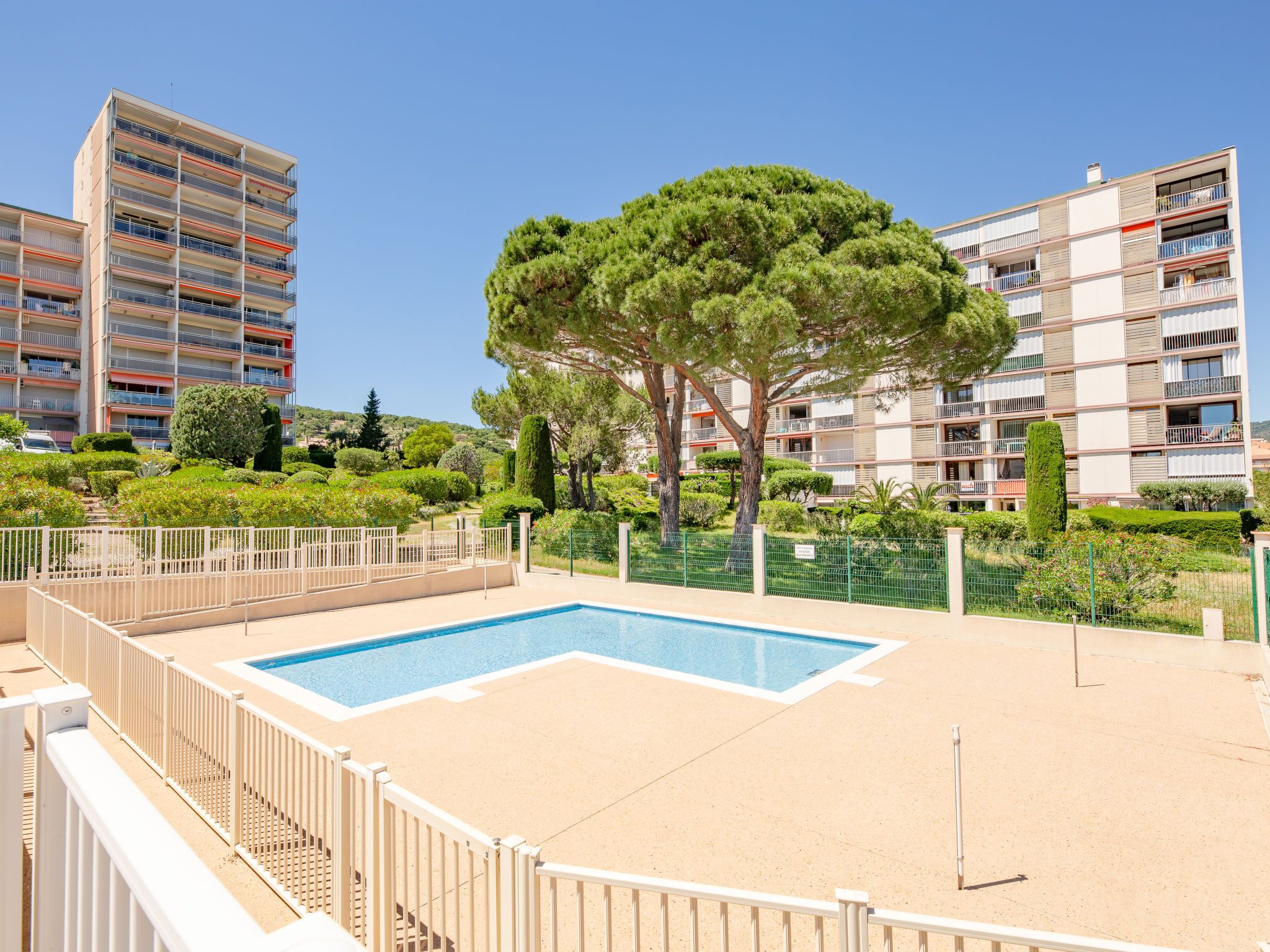 Un espace piscine doté d’une terrasse entourée de clôtures, bordé de verdure et entouré d’immeubles d’habitation.