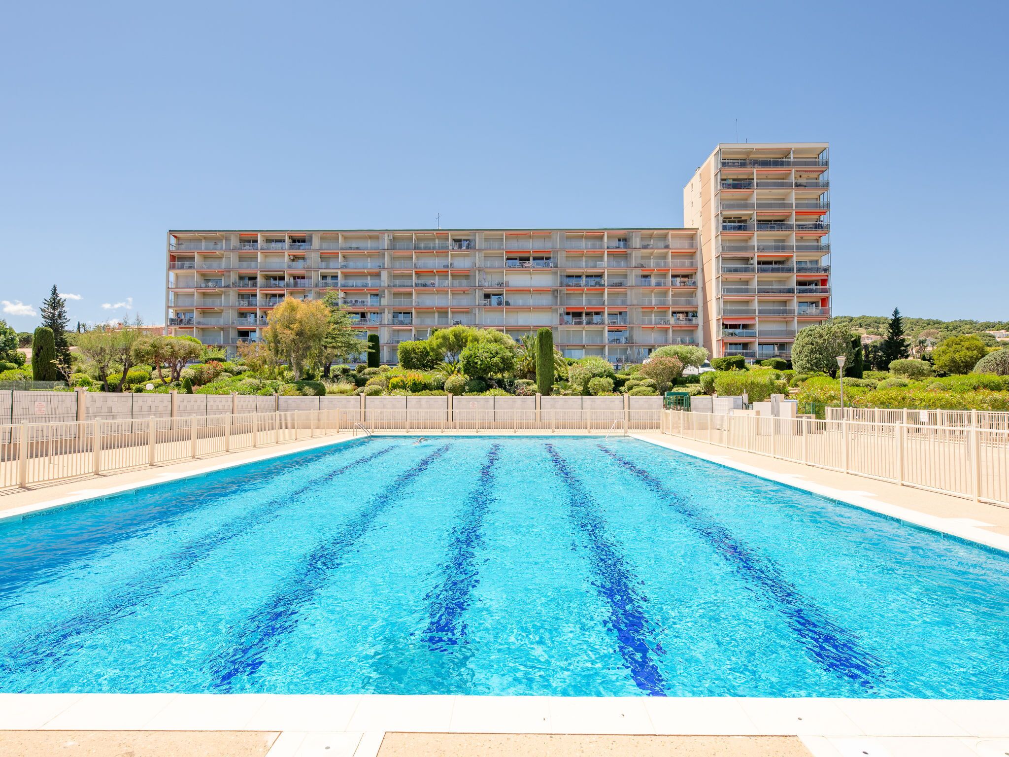 Une piscine à l’eau d’un bleu limpide, entourée d’une clôture. Au fond, on aperçoit un immeuble à plusieurs étages doté de balcons, ainsi qu’un jardin agrémenté de diverses plantes.