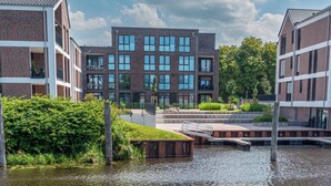 Un bâtiment moderne à la façade en briques et aux grandes fenêtres, situé à côté d’un plan d’eau, avec un quai en bois.