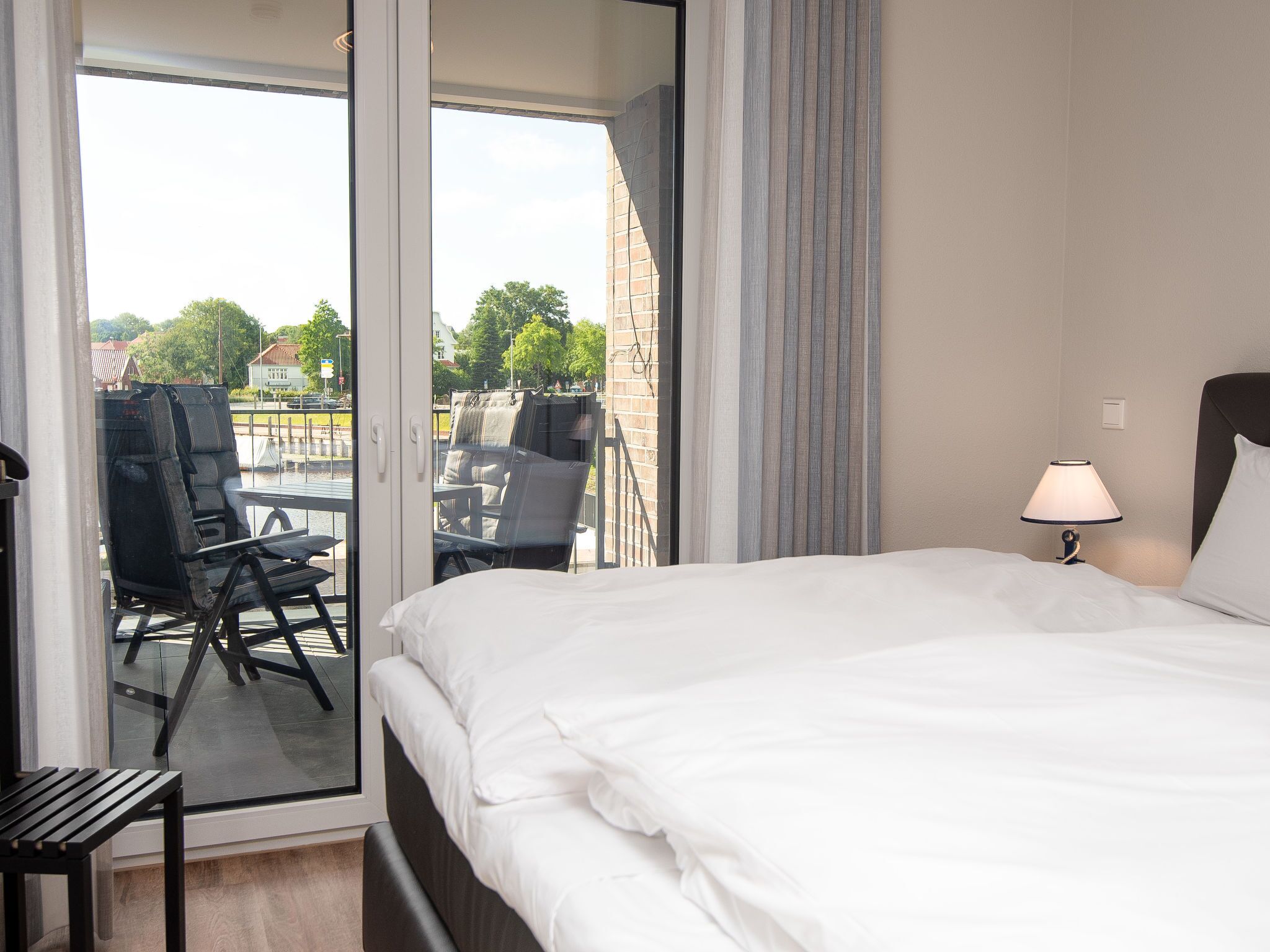 Habitación de hotel con cama, mesita de noche, lámpara y vistas al balcón exterior con sillas y una mesa.