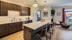A modern kitchen with dark cabinets, a large island with bar stools, pendant lighting, and a view to the living area. - Blue River Parkway’s Colorful Escape (Silverthorne)