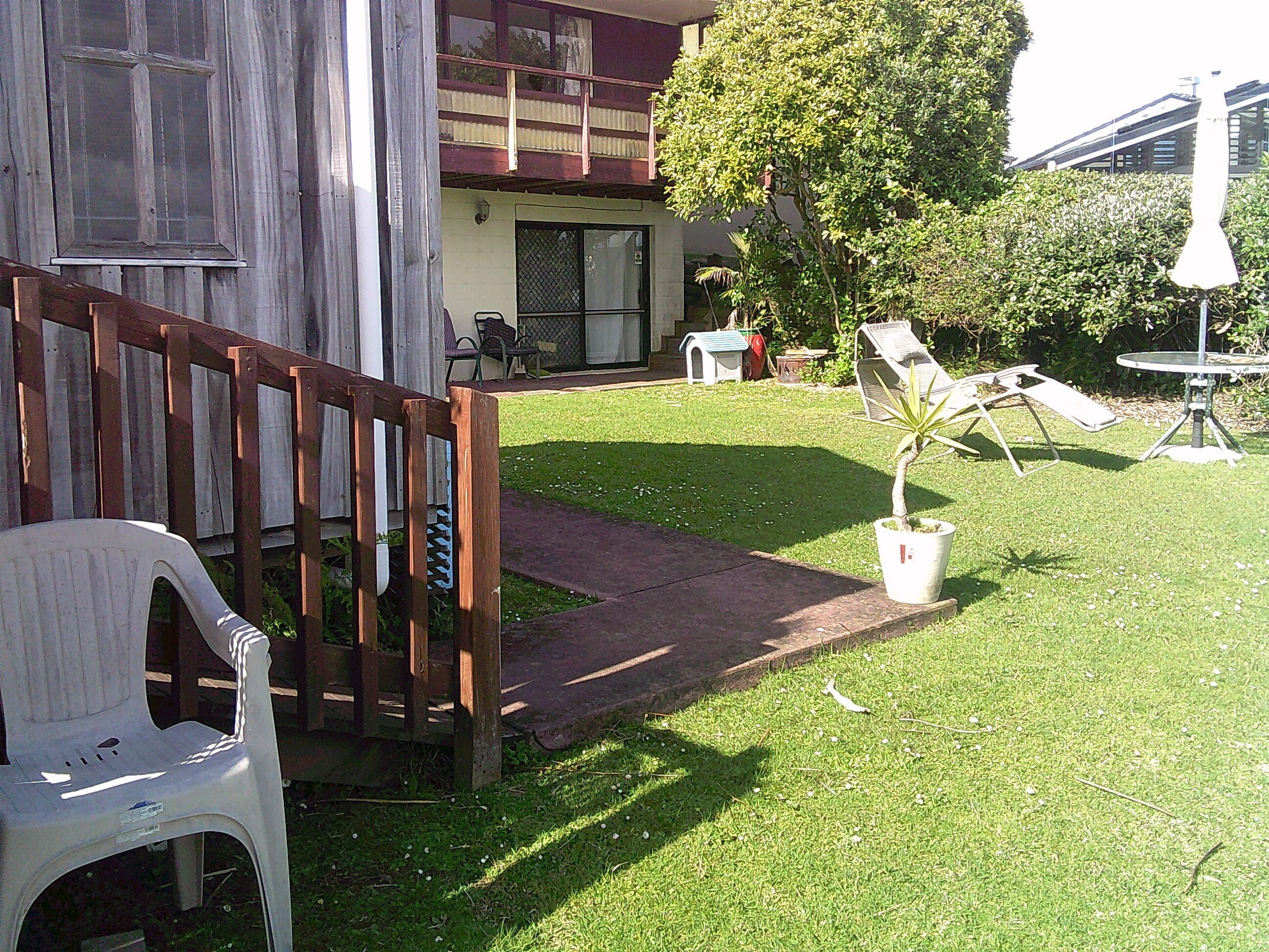 Ein Wohnhaus mit einer Holzterrasse, einem Blumentopf, einer Rasenfläche und einem kleinen Tisch mit einem Stuhl.