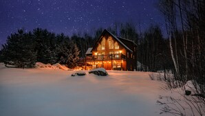Uma cabana rústica com varanda, rodeada por terreno e árvores cobertas de neve, sob um céu estrelado.