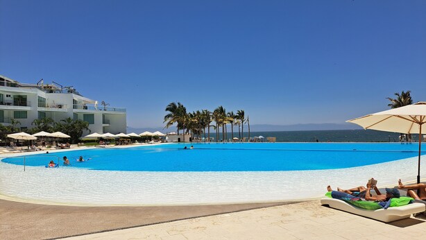 A large swimming pool with a clear blue sky, palm trees, and a beach area with lounge chairs and umbrellas. - Aria Ocean Nuevo Vallarta Beach Pool (Nuevo Nayarit)