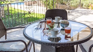 Taula rodona de vidre amb cadires de vimet, una tassa blanca, un got de beguda vermella i un bol de llaminatures en un balcó amb vista a un jardí i una piscina.