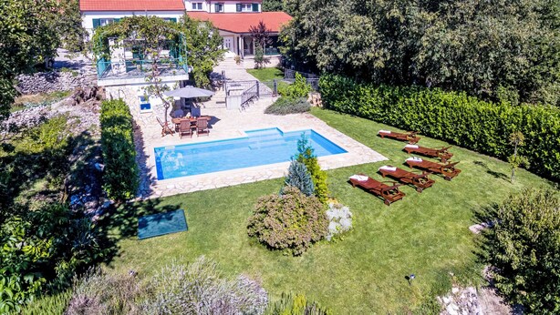Una piscina envoltada d'un jardí ben cuidat, un porxo amb mobles d'exterior, i una casa amb teulada vermella al fons.