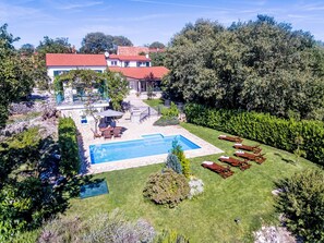 A swimming pool surrounded by a well-maintained lawn, a patio with outdoor furniture, and a house with a red roof in the background.