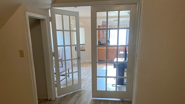 A room with a wooden floor, a bed with a white comforter, and a door leading to a kitchen area. - Cozy Amazing Suite  (Willmar)