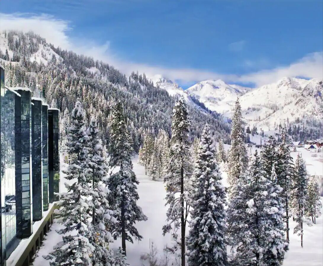 A snow-covered landscape with a row of modern buildings, likely a hotel, with a view of snow-laden trees and mountains in the background.