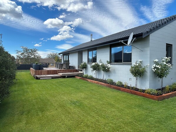 A modern house with a well-maintained lawn, a patio with outdoor furniture, and white flowering plants along the side. - Elevated Mountain Views on Waenga (Cromwell)