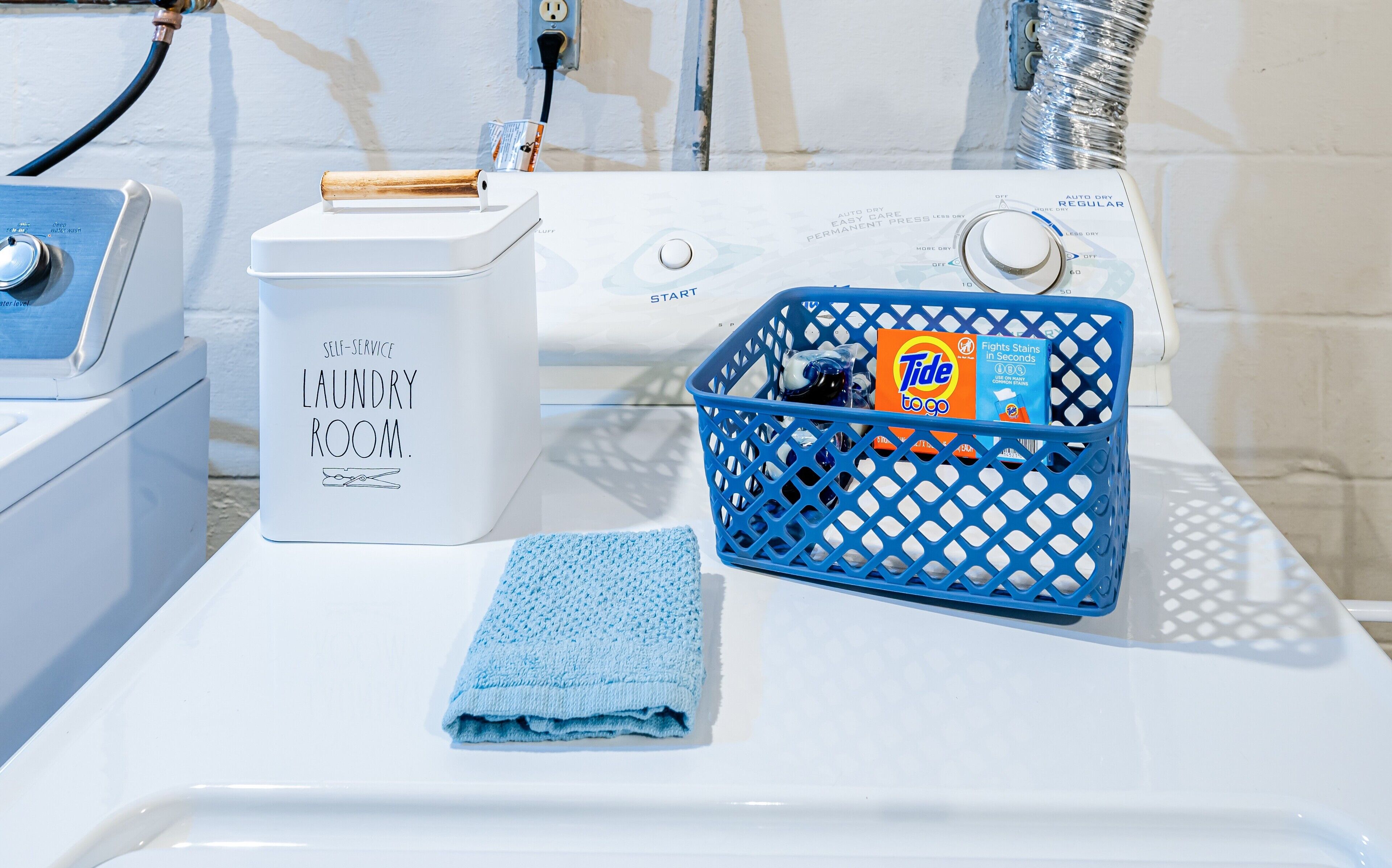 Een wasserette met een wasmachine, een droger en een blauwe wasmand met Tide wasmiddel.