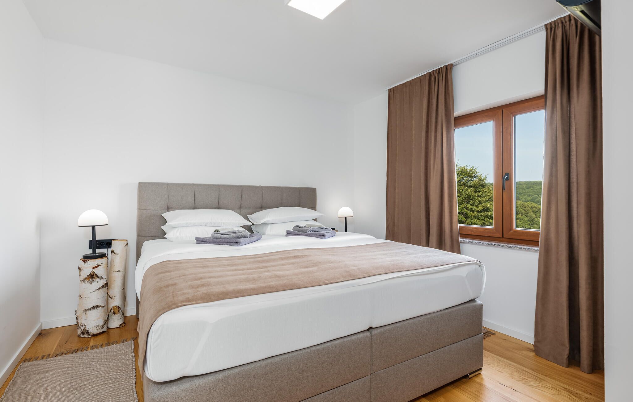 A bedroom with a bed, two bedside lamps, a window with curtains, and a view of greenery outside.