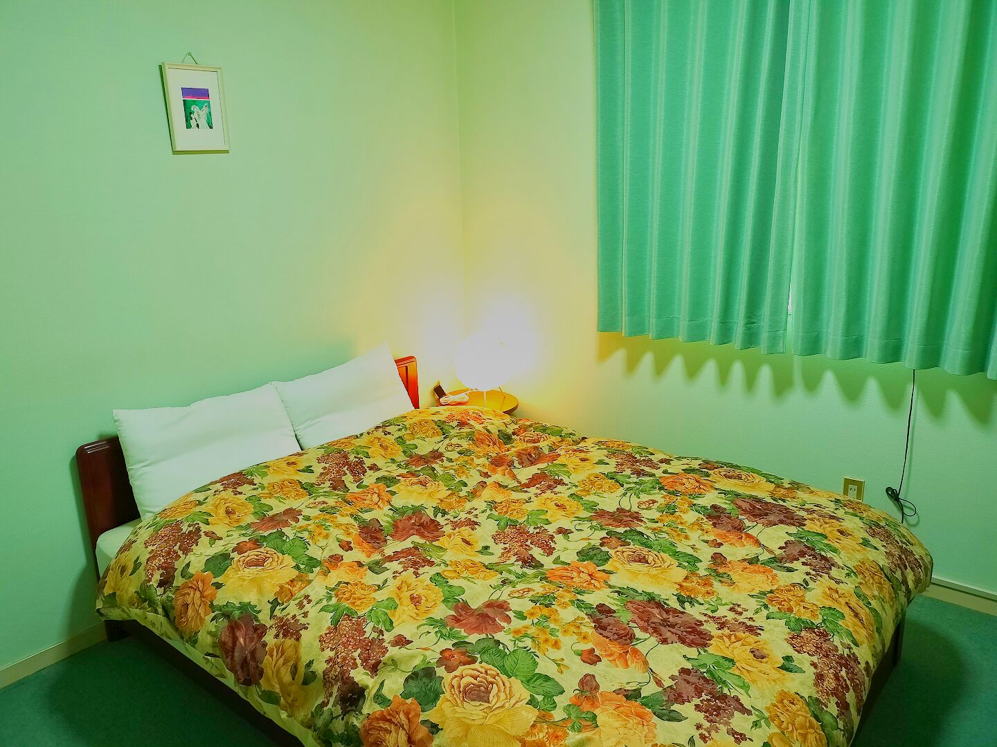A room with a bed, a bedside table, a lamp, a framed picture on the wall, and a window with curtains.
