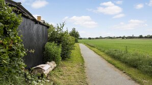 Un sentier rural mène à un bâtiment en bois doté d’une cheminée, entouré de verdure et de champs.