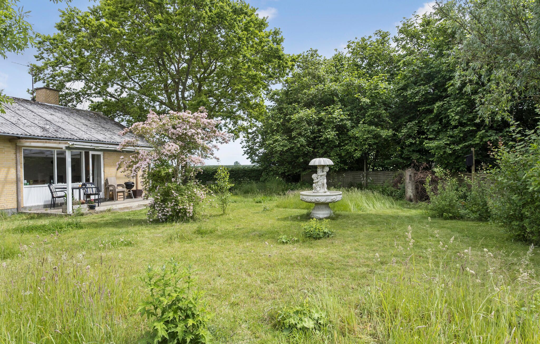 Ein Garten mit einer Fontäne, eine Terrasse mit Stühlen und ein Haus mit Satteldach.