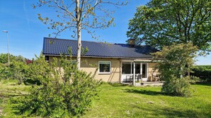 Une maison de plain-pied avec un toit recouvert de tuiles bleu foncé, un porche à l’avant avec une table et des chaises, et un petit jardin agrémenté de divers arbustes et d’un arbre.
