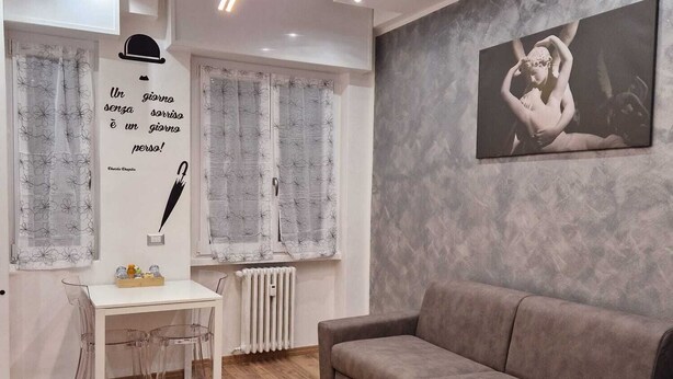 A modern living room with a grey sofa, a white table with chairs, and a radiator. There is a quote on the wall and a framed picture above the sofa. - Canova Accogliente Casa Vacanze - Affari (Milano)