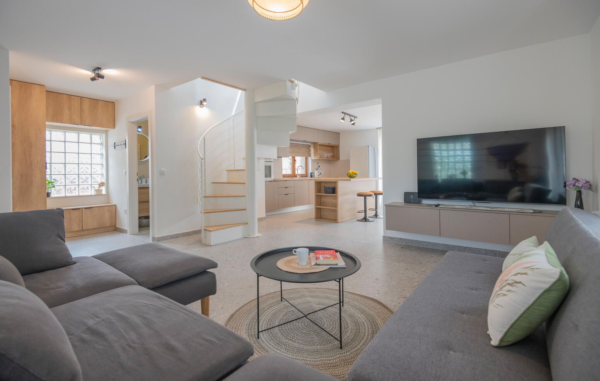 A modern living room with a sofa, a coffee table, and a TV. There is a kitchen area with a staircase leading to another level.