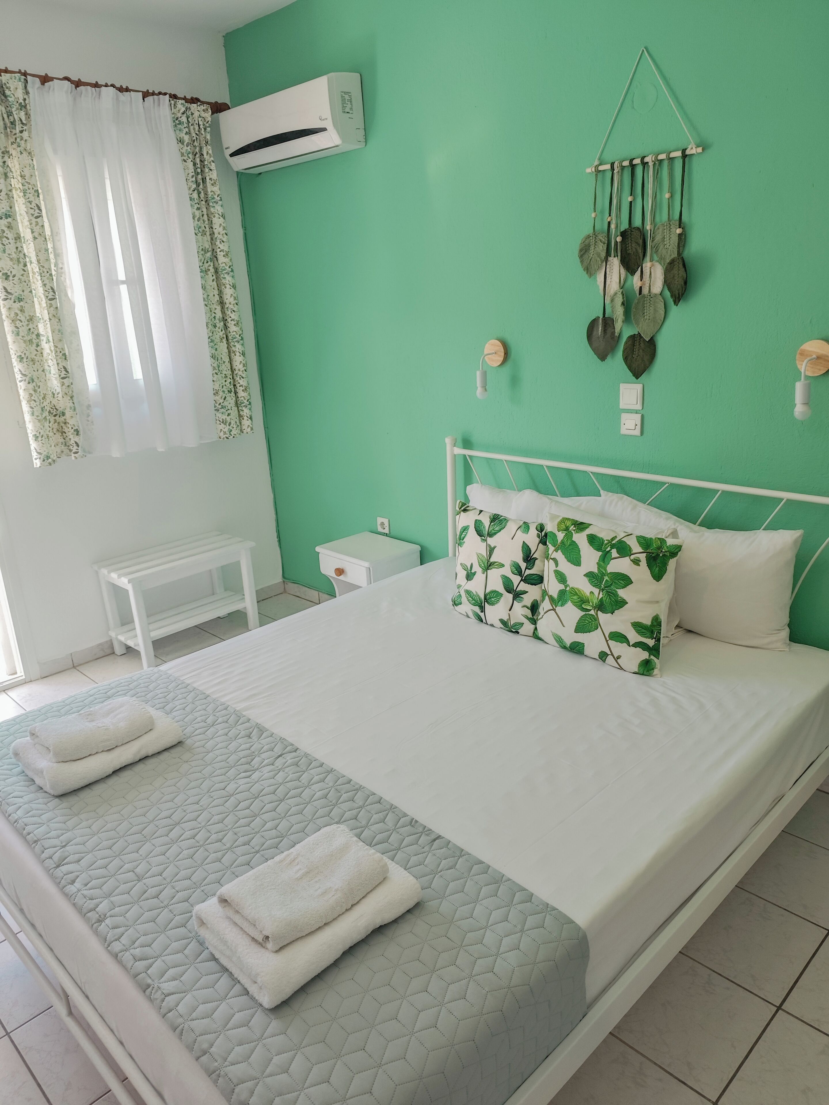 A neatly made bed with white bedding and a green leaf-patterned pillow, a bedside table, a small nightstand, and a wall-mounted air conditioner.