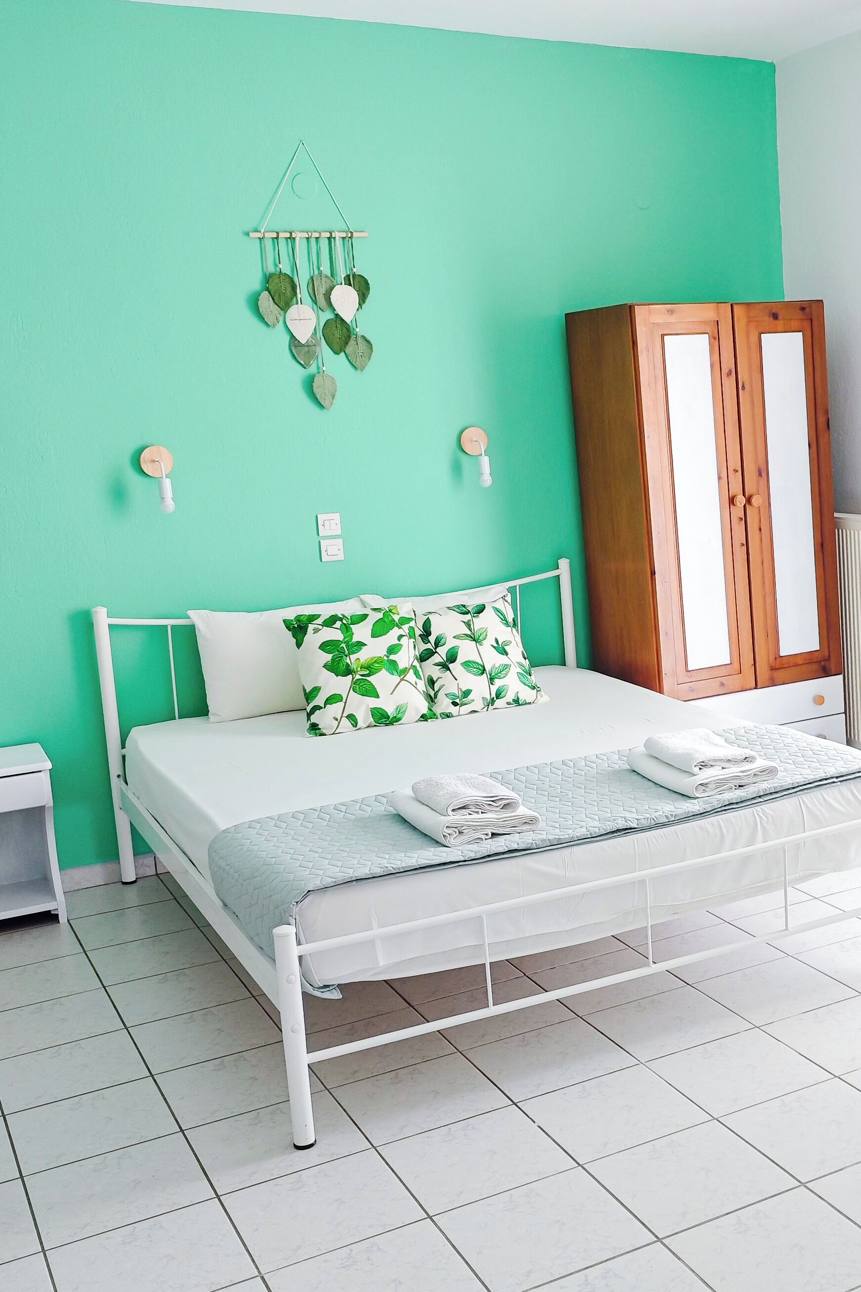Un lit bien fait, avec une literie blanche et un coussin orné d’un motif de feuilles vertes, une armoire en bois et un luminaire fixé au mur.