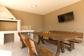 A dining area with a large wooden table, benches, a brick oven, and a television mounted on the wall.