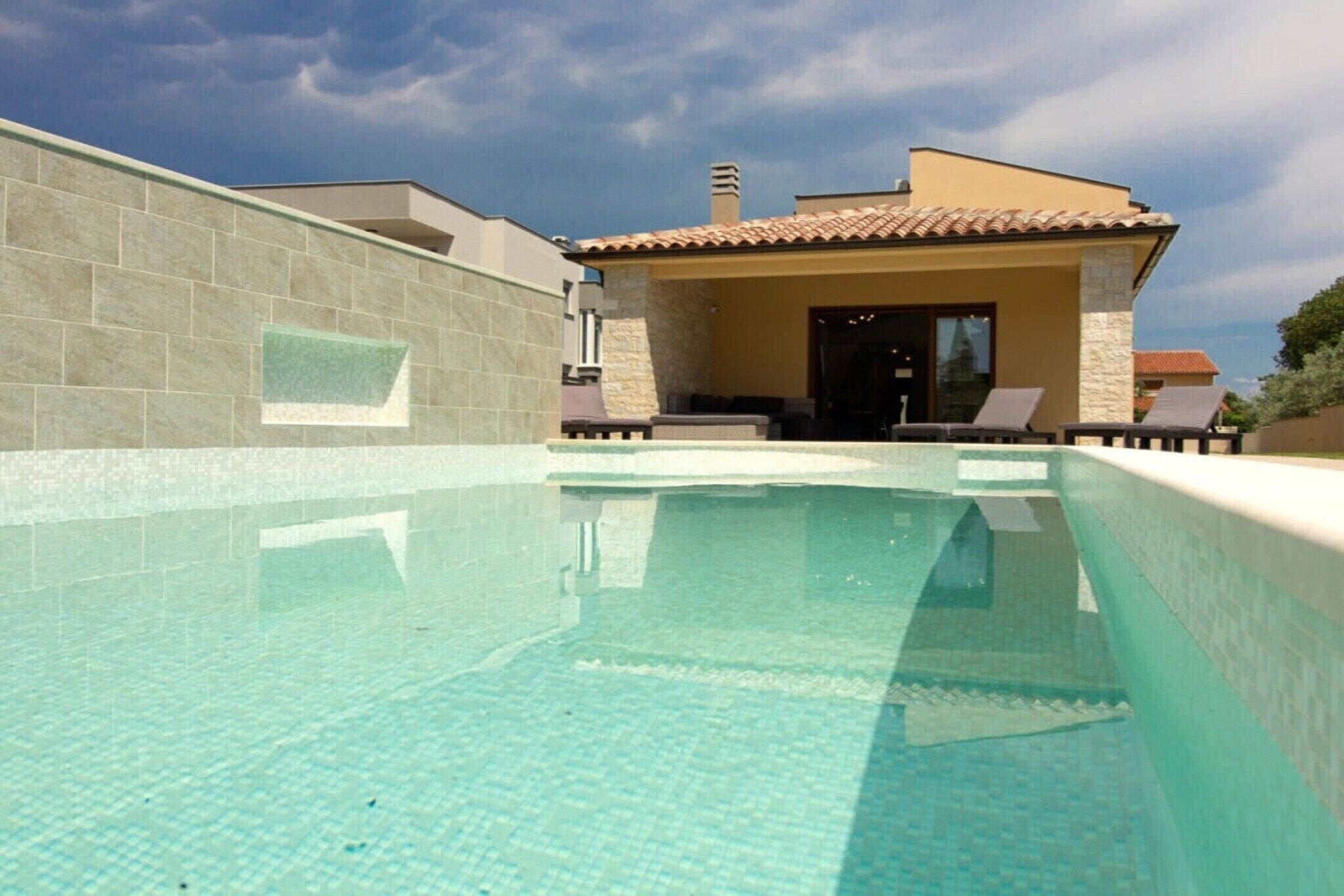Une piscine à l’eau claire, un patio couvert avec des chaises longues et un bâtiment doté d’un toit en tuiles.