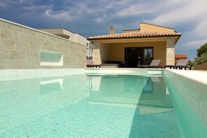 A swimming pool with clear water, a covered patio area with lounge chairs, and a building with a tiled roof.