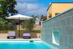 Une piscine avec des chaises longues et un parasol, une tour en pierre en arrière-plan et un bâtiment à la façade recouverte de carreaux.