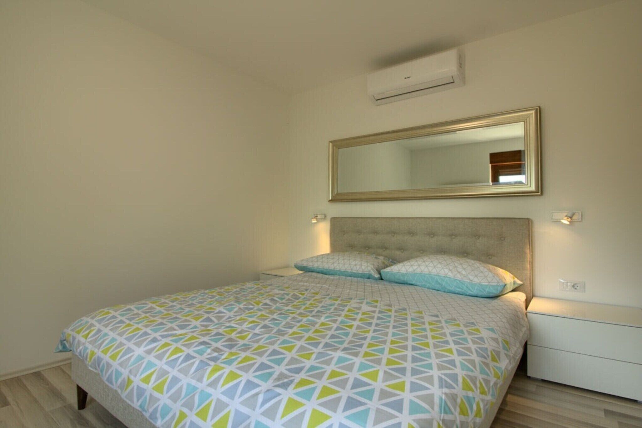 A bedroom with a bed, a headboard, a wall-mounted mirror, and a bedside table.
