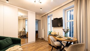 Een moderne woonkamer met een groene bank, een houten eettafel met stoelen, een aan de muur bevestigde televisie en een groot raam met gordijnen.