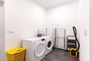 A laundry room with a front-loading washing machine, a drying rack, a vacuum cleaner, and a yellow laundry basket.