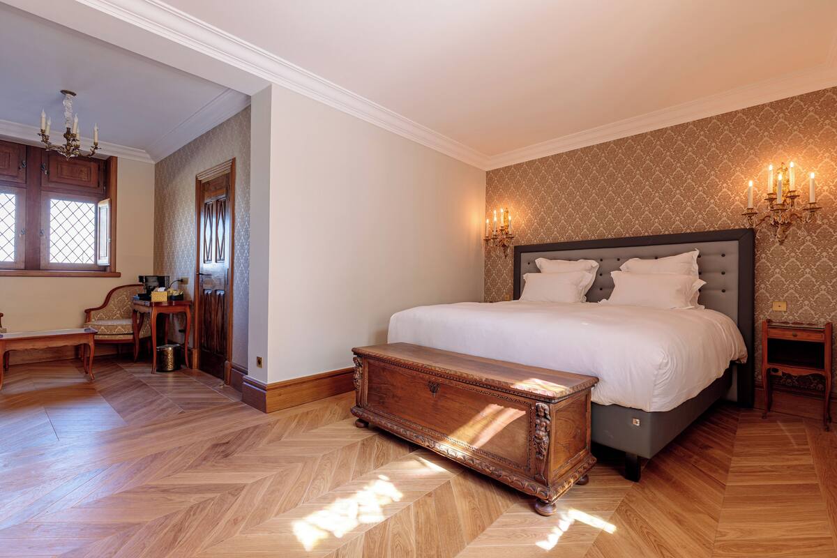 A bedroom with a large bed, wooden floor, a bench, a chest of drawers, and a chandelier.