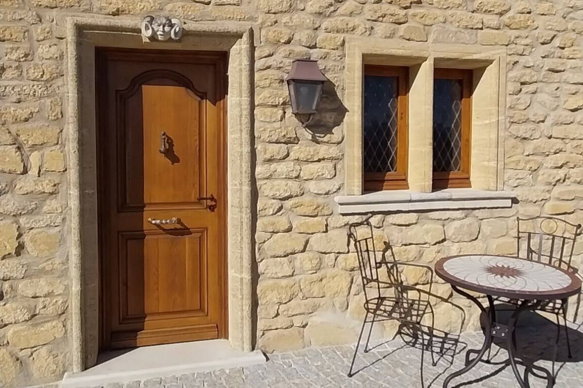 A stone building with a wooden door, a small window, and a table with chairs outside.