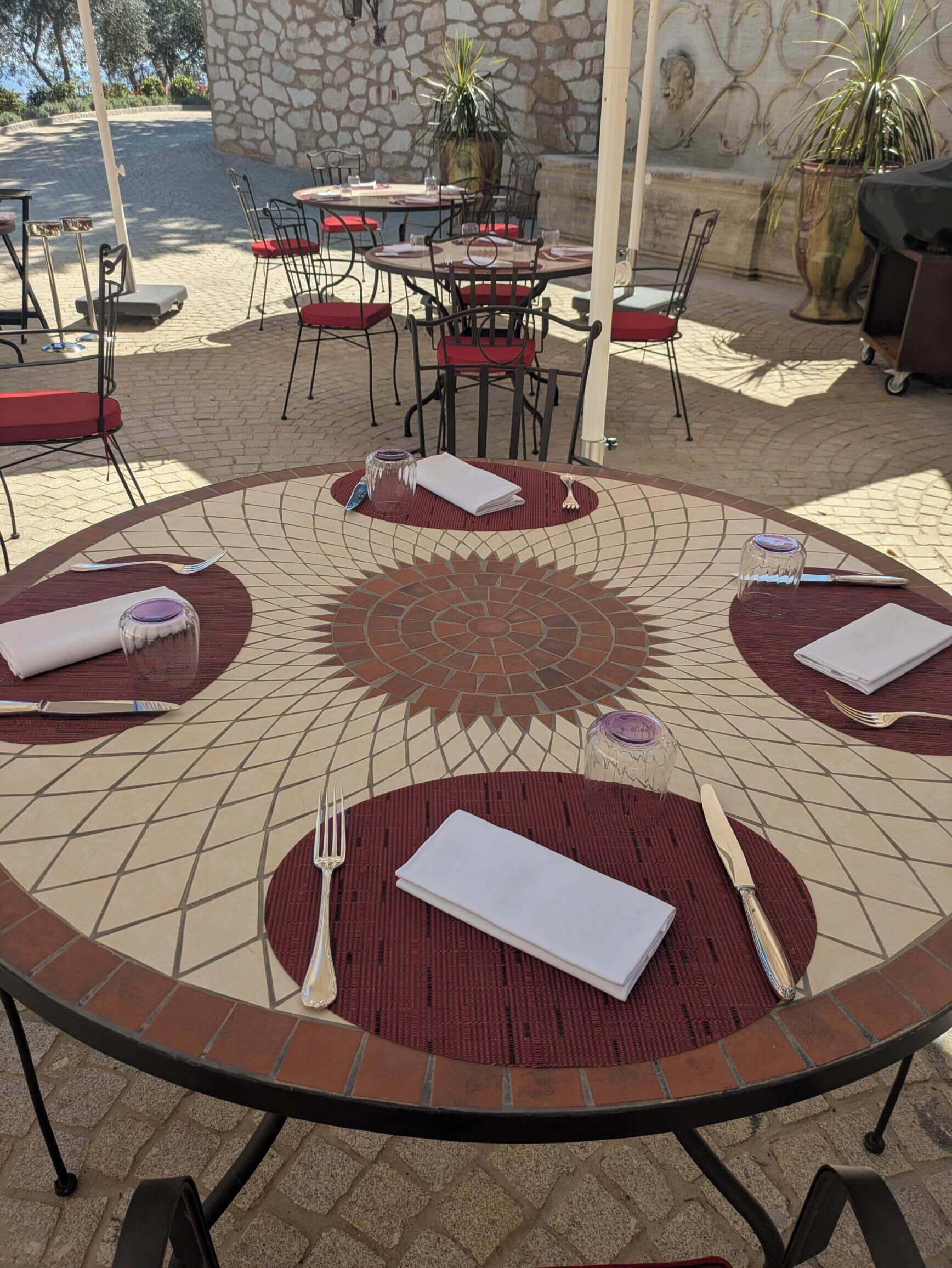 A round outdoor dining area with red and beige mosaic tables, black metal chairs, and a stone wall backdrop.