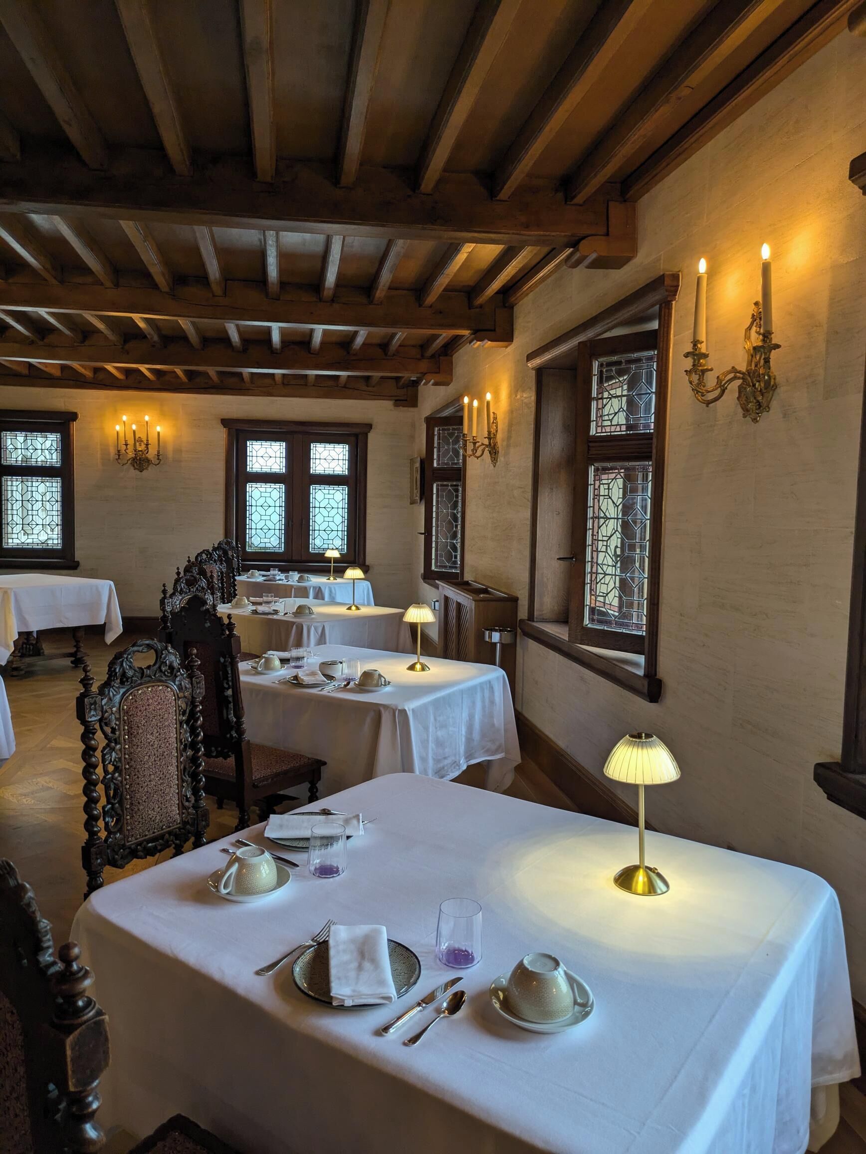Uma sala de jantar com mesas preparadas para quatro pessoas, vigas de madeira no teto e janelas de vitrais.