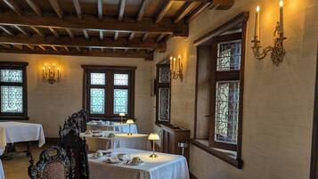 Uma sala de jantar com mesas preparadas para quatro pessoas, vigas de madeira no teto e janelas de vitrais.