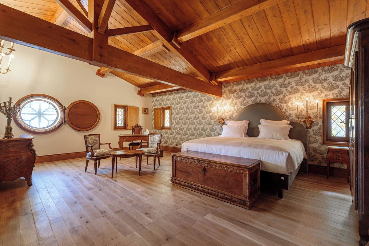 A spacious bedroom with a large bed, wooden ceiling beams, and a stone wall.