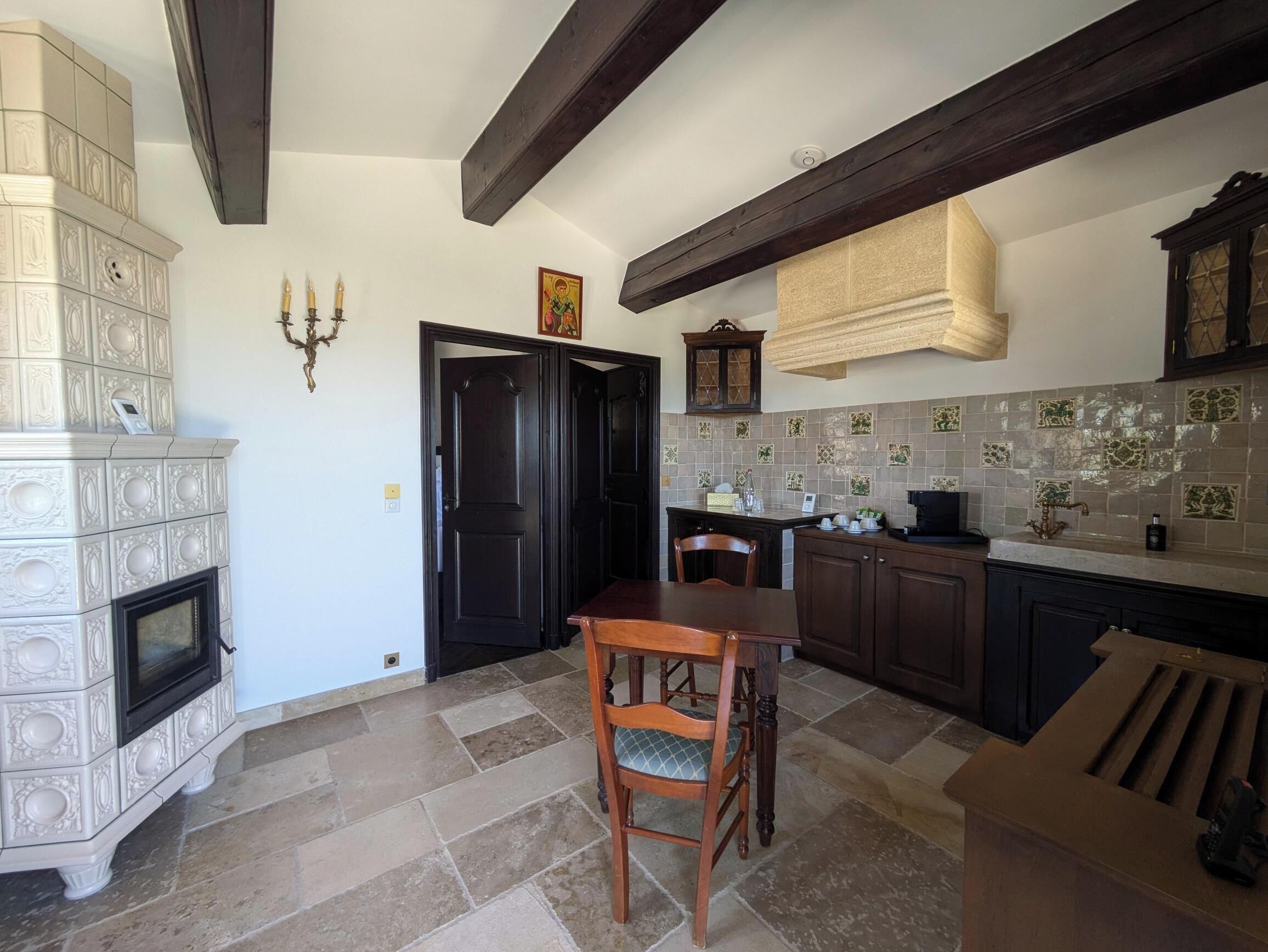 Una cocina con chimenea, vigas de madera oscura, salpicadero de azulejos y un comedor con mesa y sillas de madera.
