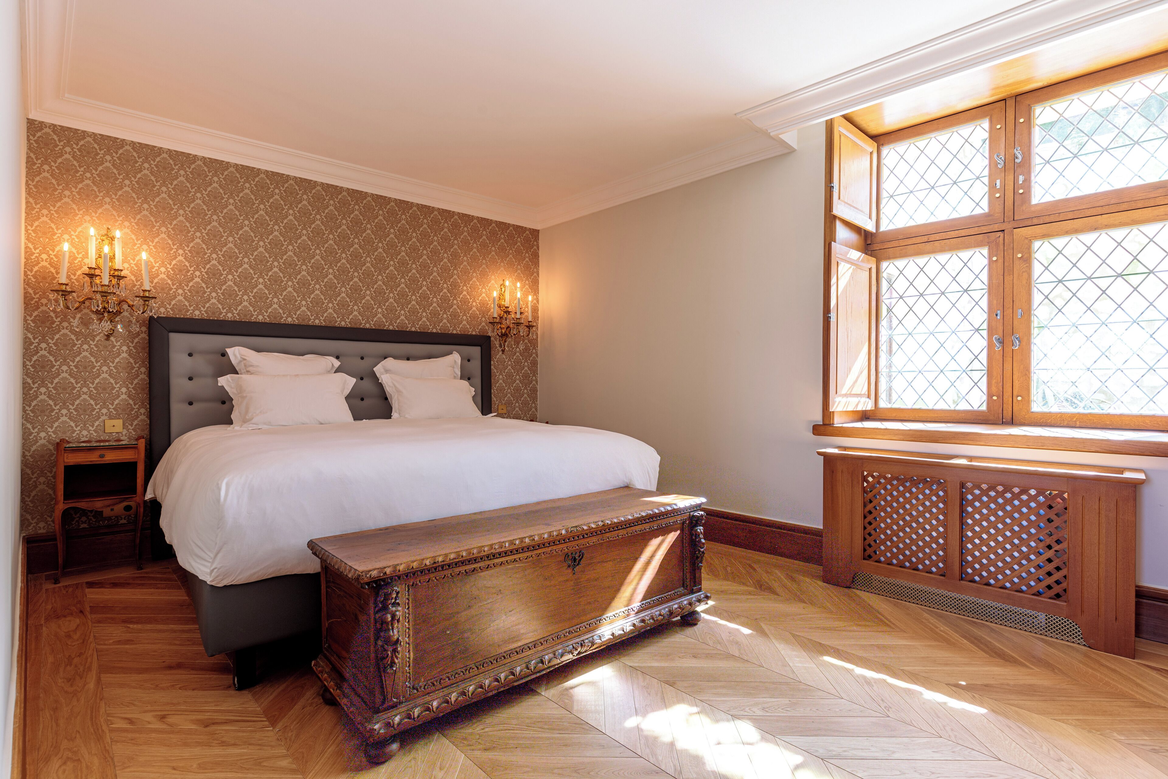 A bedroom with a large bed, a wooden chest, two wall sconces, and a window with a wooden frame.