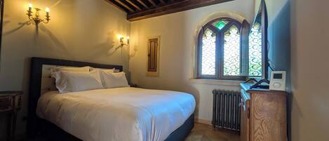 A bedroom with a bed, a wooden headboard, a nightstand, a wall-mounted lamp, a radiator, a wooden dresser, and a window with stained glass.