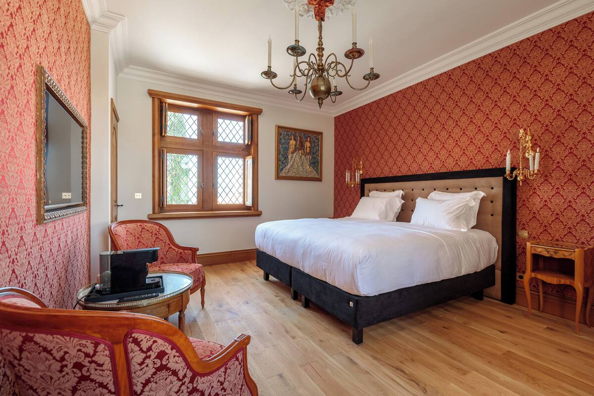 A bedroom with a large bed, a chandelier, a wooden floor, and patterned wallpaper.
