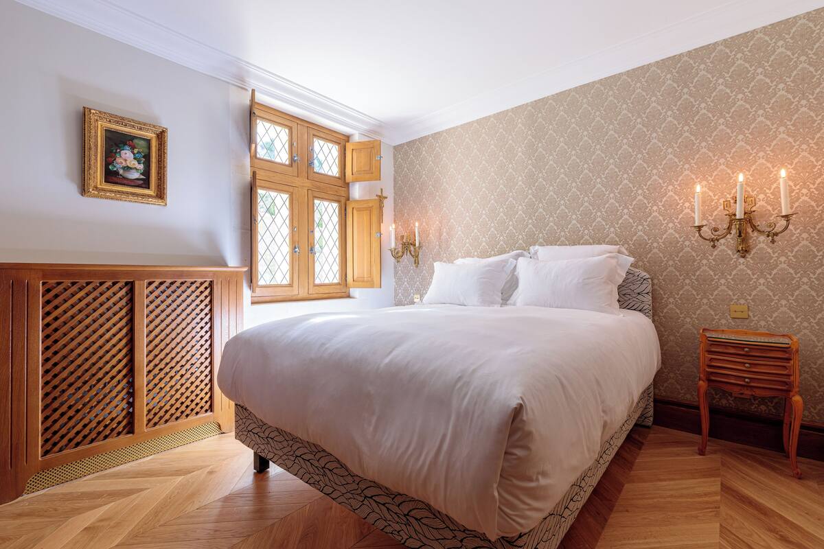 A bedroom with a large bed, wooden floor, a window with a view, a wall-mounted picture, and a bedside table with candles.