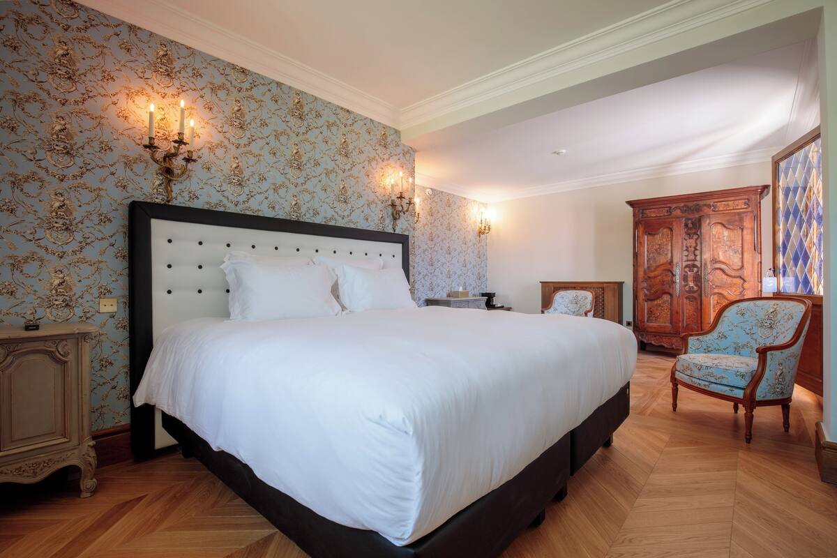 A large bed with a tufted headboard, two bedside tables, a wooden wardrobe, and a patterned chair.
