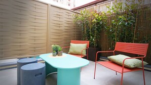 Un espace extérieur moderne, aménagé avec une table turquoise et des chaises en métal rouge, entouré de plantes en pot et d’une clôture en bambou.