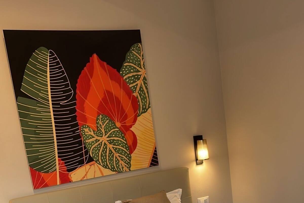 Une chambre à coucher avec un grand lit, une tête de lit, une lampe fixée au mur et une œuvre d’art colorée au-dessus du lit.