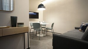 Un salon moderne avec un canapé gris, une table basse et un téléviseur fixé au mur. On y trouve également un coin repas avec une table ronde et quatre chaises.