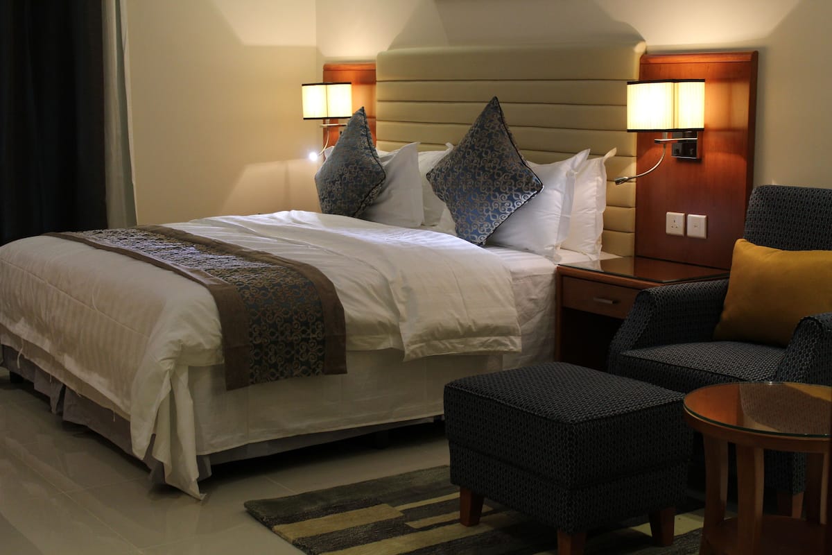 A hotel room with a bed, bedside tables, a chair, a small table, and a lamp.