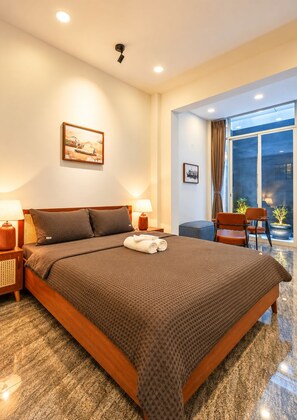 A hotel room with a large bed, two bedside lamps, a window with curtains, a small table with chairs, and a painting on the wall. - Nexus House Cay Diep - Near Independence Palace (Ho Chi Minh City)